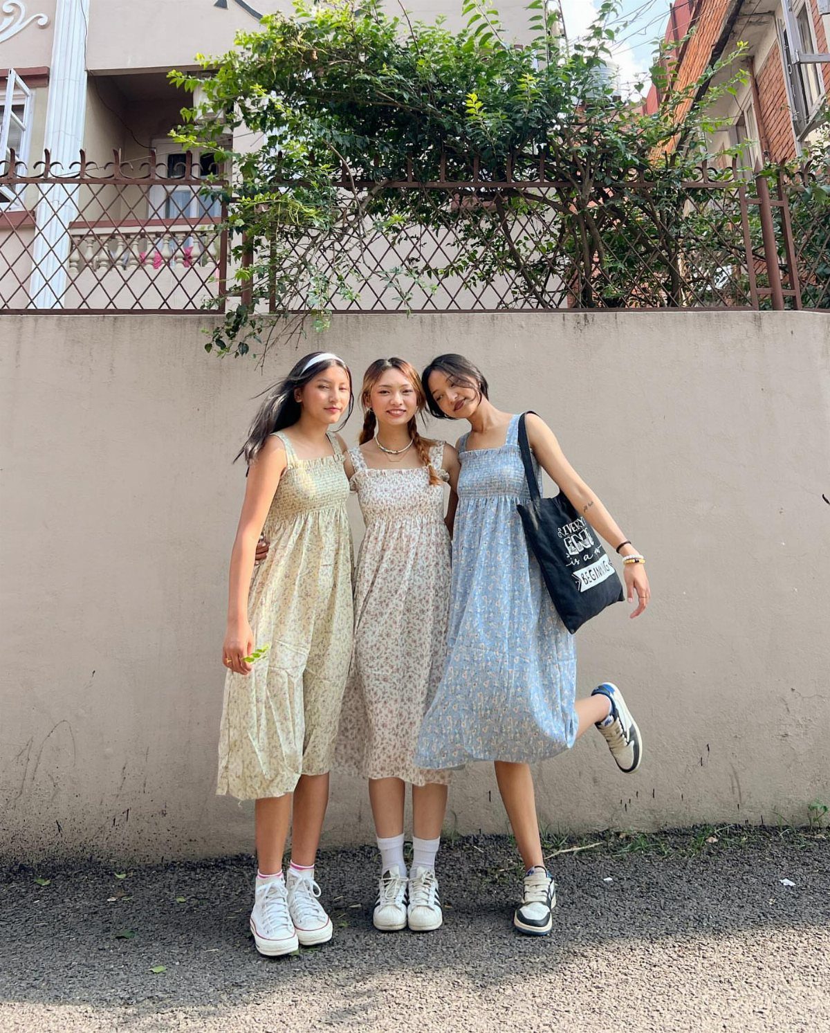 Three girls posing together wearing floral sundresses and white sneakers against a wall.