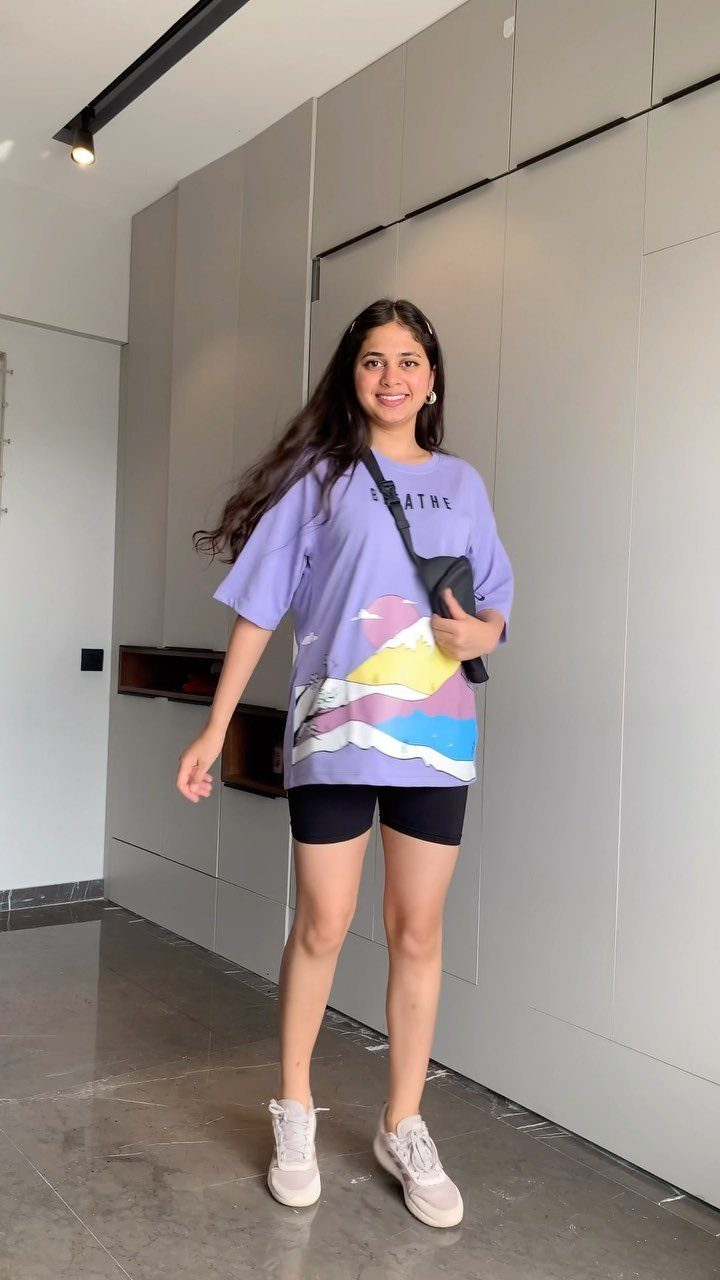 A smiling young woman wearing an oversized lilac graphic tee and black biker shorts.
