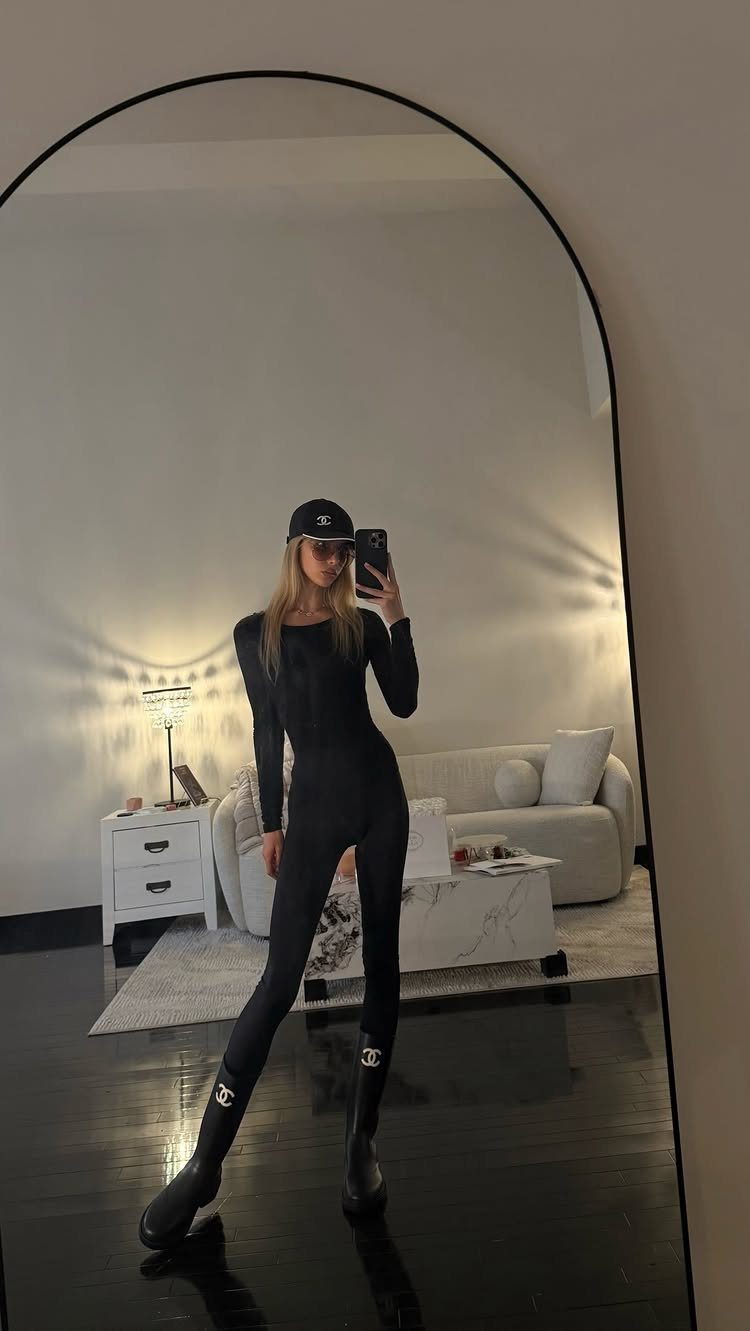 Woman in a black bodysuit and cap posing for a mirror selfie in a living room.