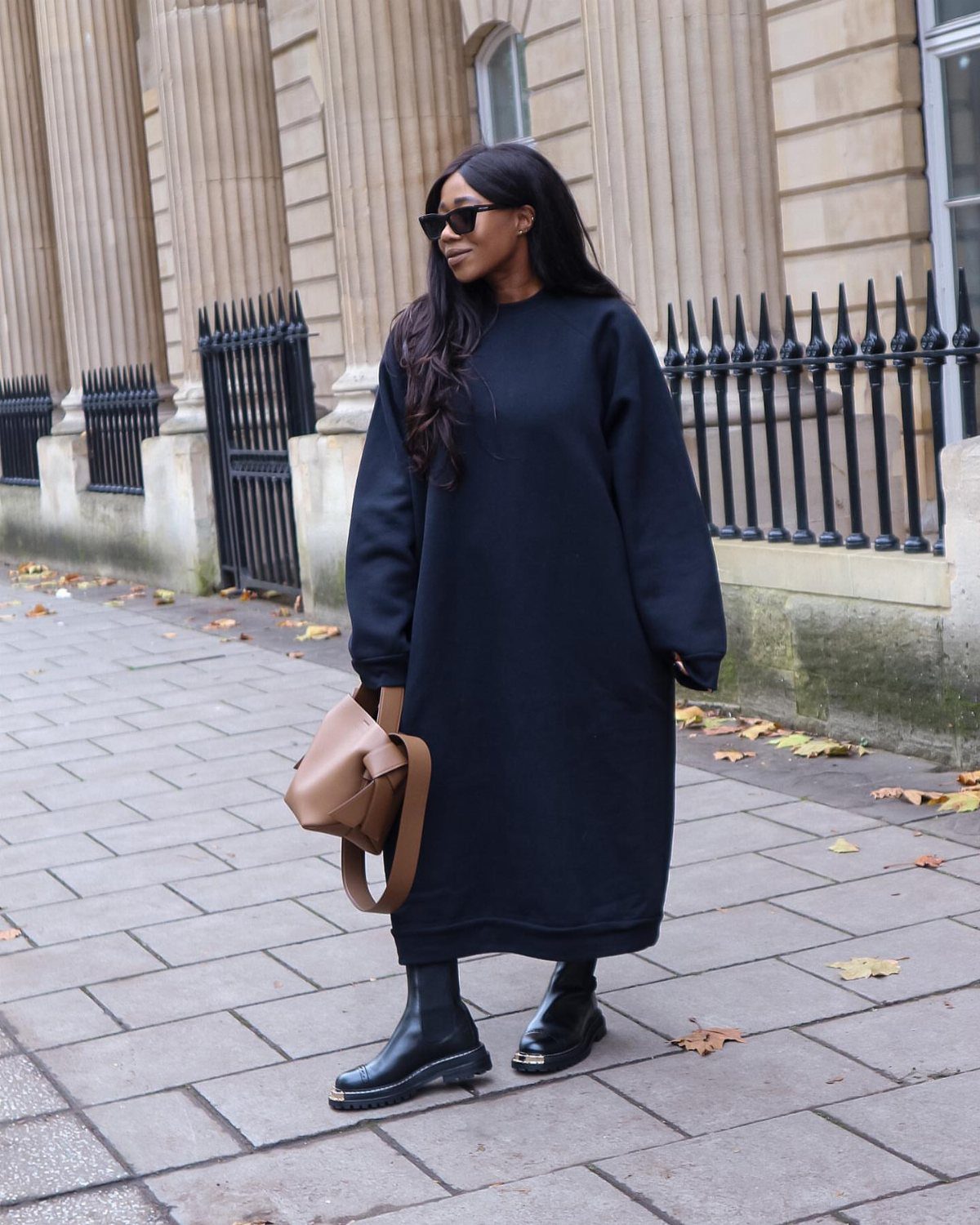 Woman in an oversized black sweater dress, sunglasses, and chunky boots standing on a city sidewalk.
