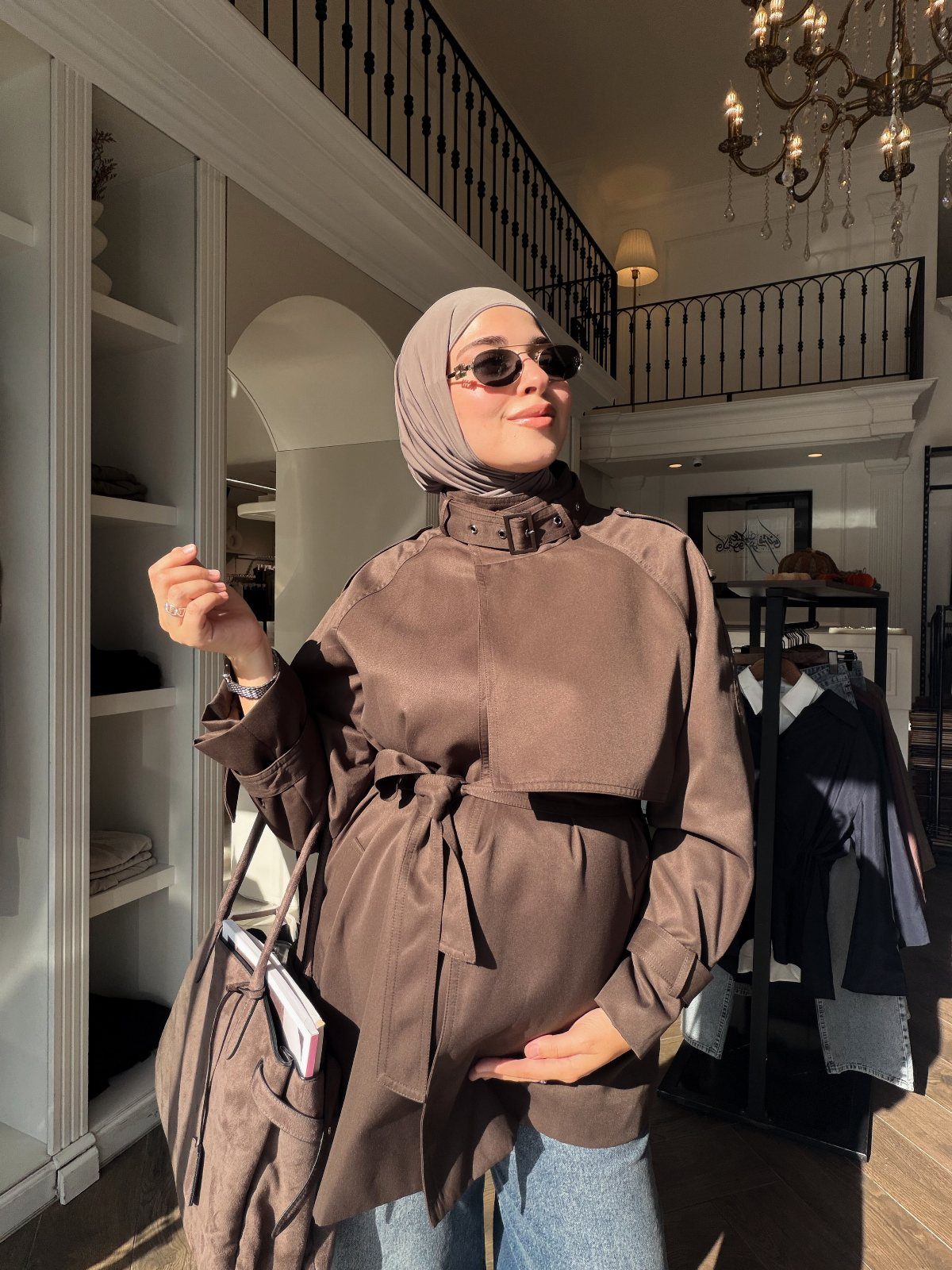 A woman in a brown belted jacket and hijab smiling in a bright clothing boutique.