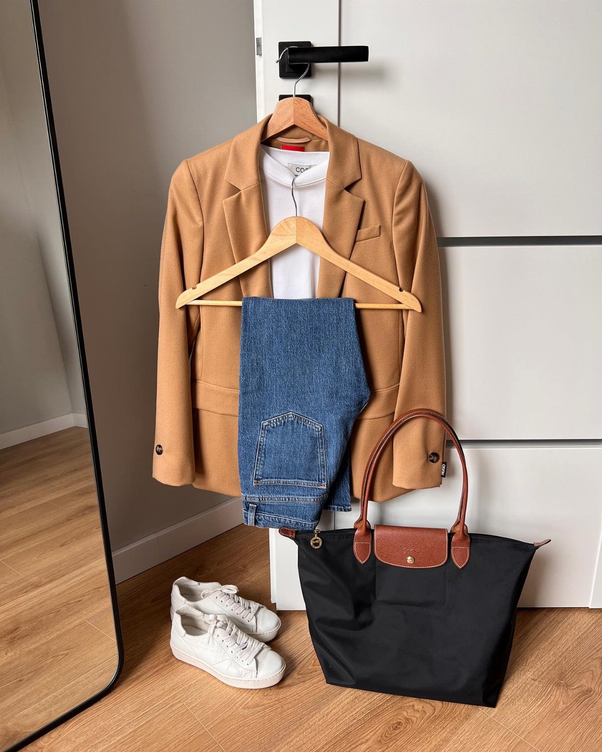 A camel blazer, white shirt, and blue jeans hanging on a door above a black tote bag.