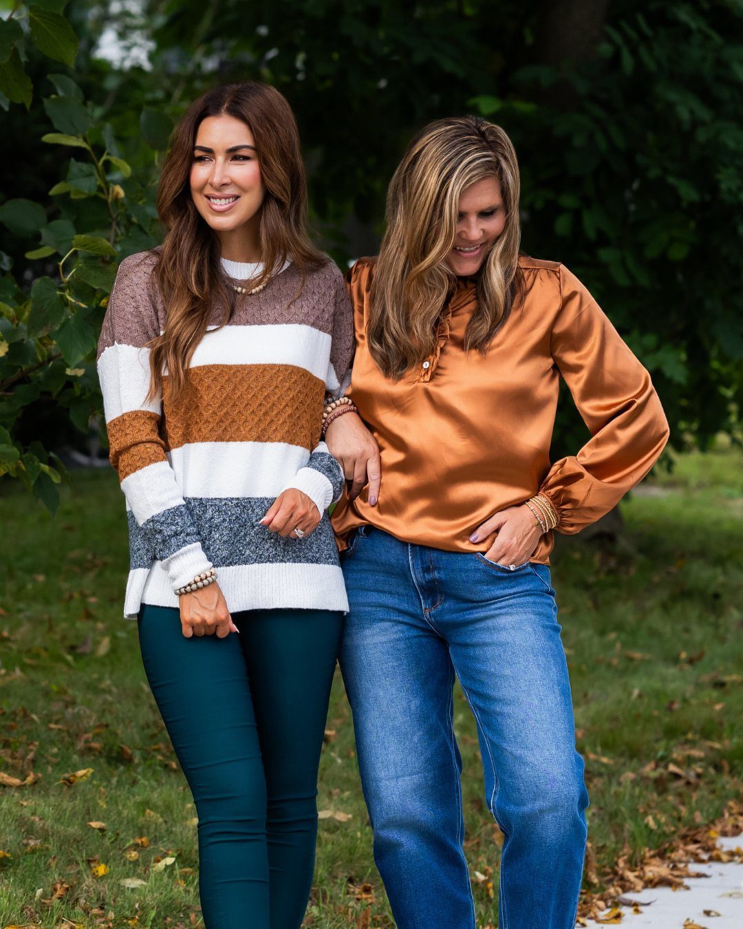 Two women walking through a park wearing a striped sweater and a copper satin blouse.