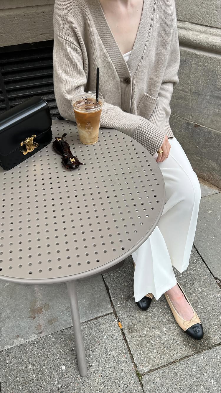 A person wearing a beige cardigan and white trousers sitting at an outdoor cafe table.