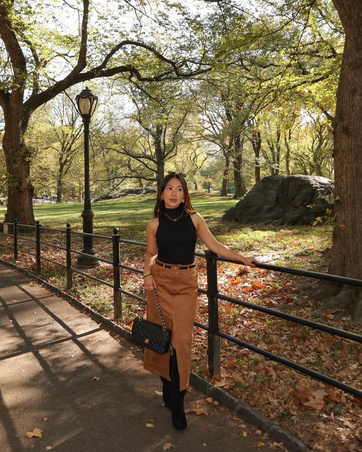 A woman in a black turtleneck and brown midi skirt posing by a park fence.