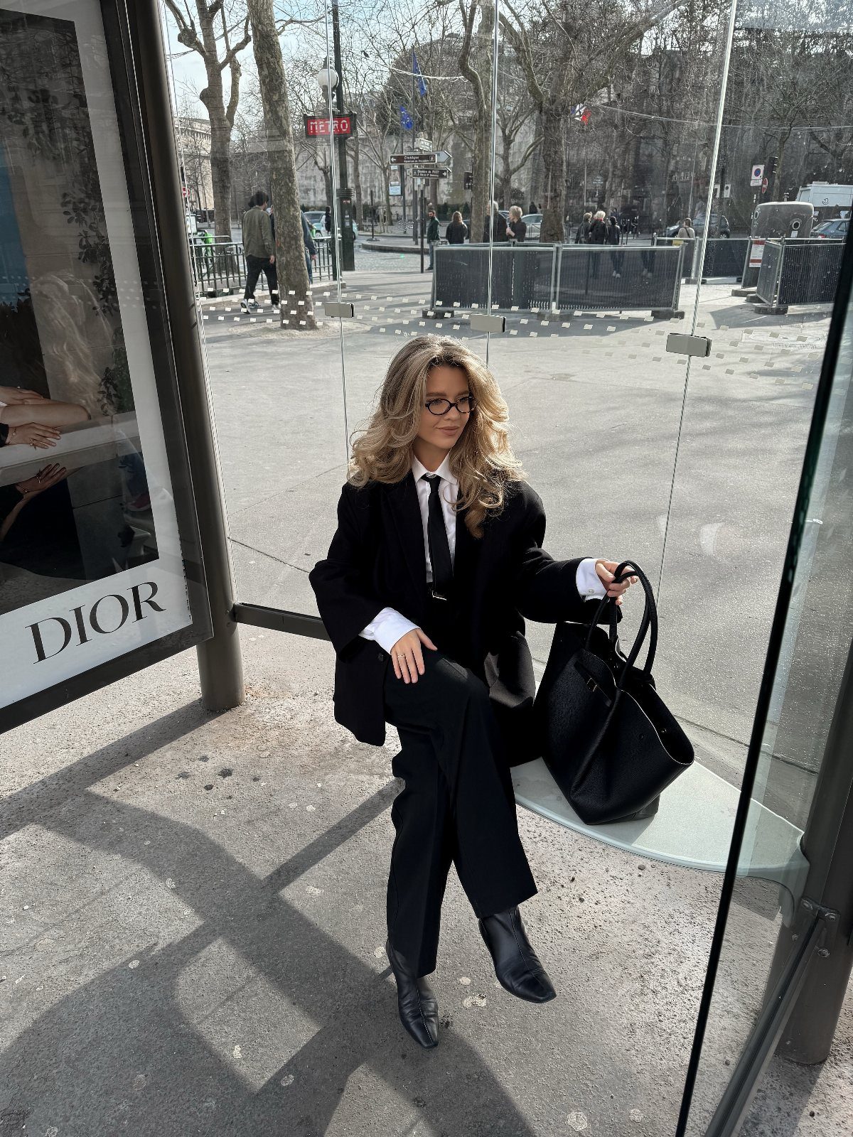 Woman in a black suit and tie sitting outdoors holding a large black handbag.