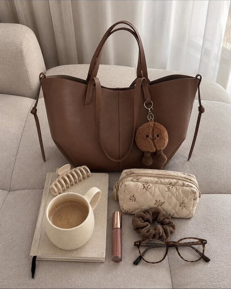 Brown leather tote bag surrounded by lifestyle accessories on a light sofa.