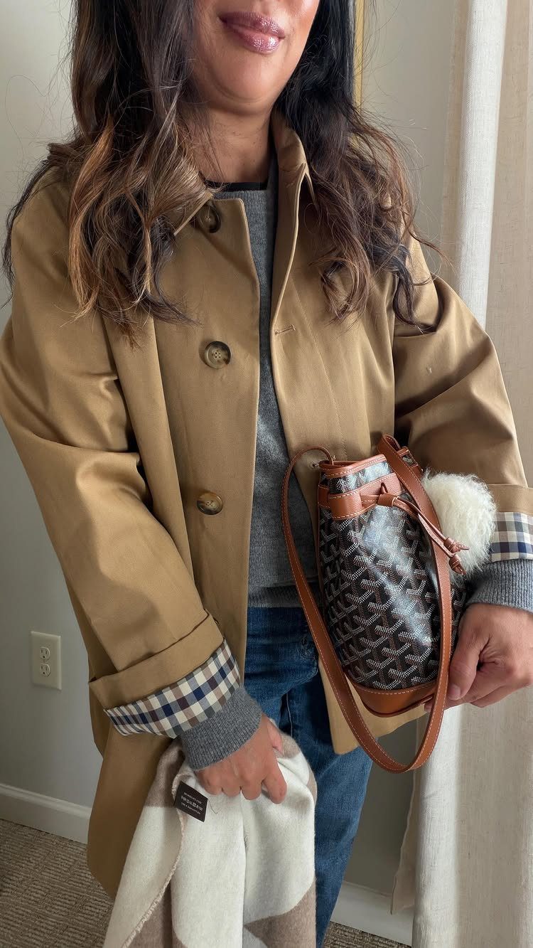 Woman wearing a tan trench coat, grey sweater, and blue jeans while holding a patterned bag.