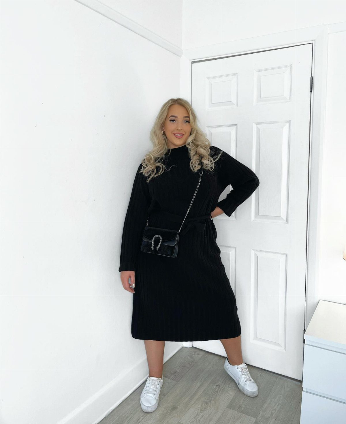 A blonde woman wearing a black midi dress and white sneakers standing indoors.
