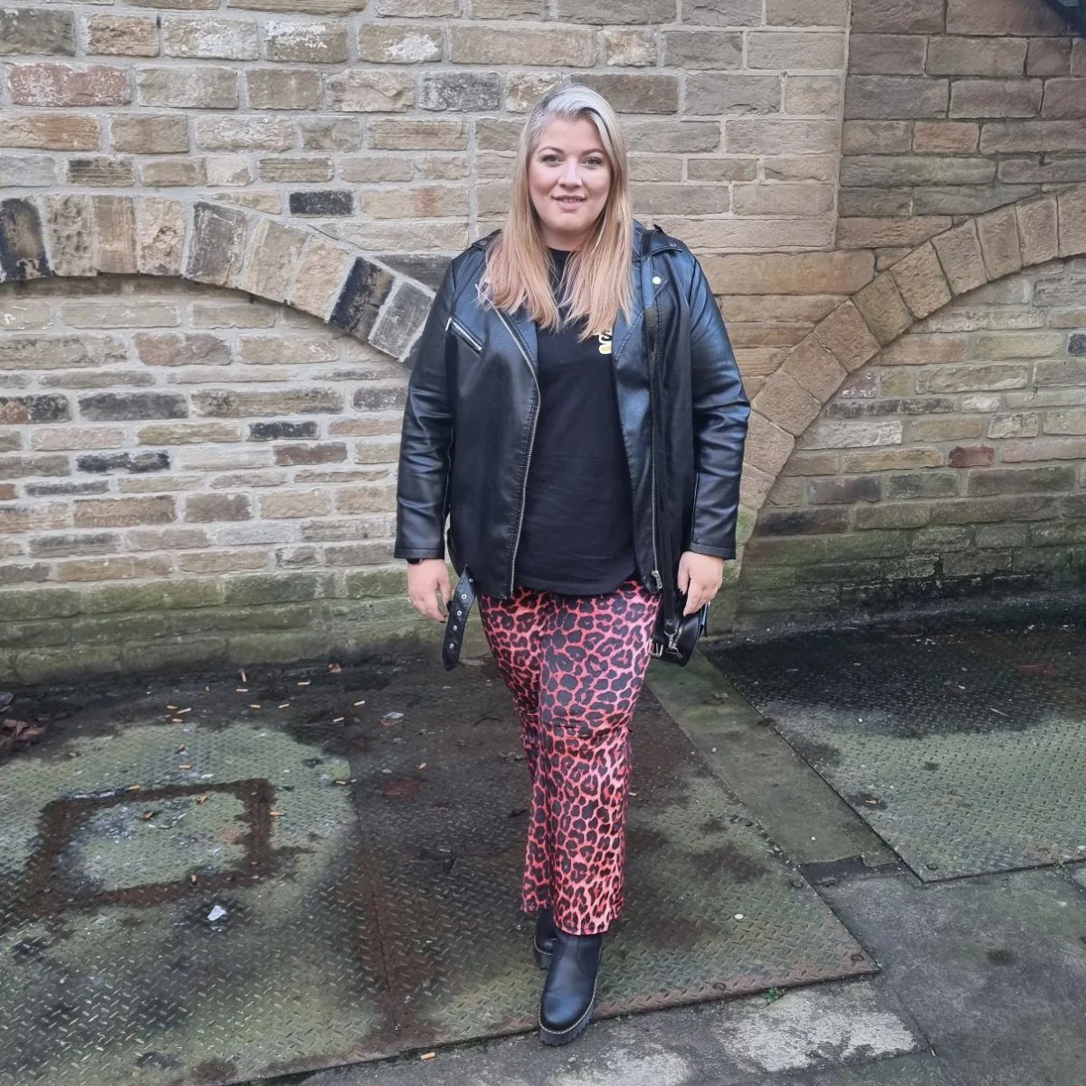 A blonde woman posing in a leather jacket and leopard print trousers against a brick wall.