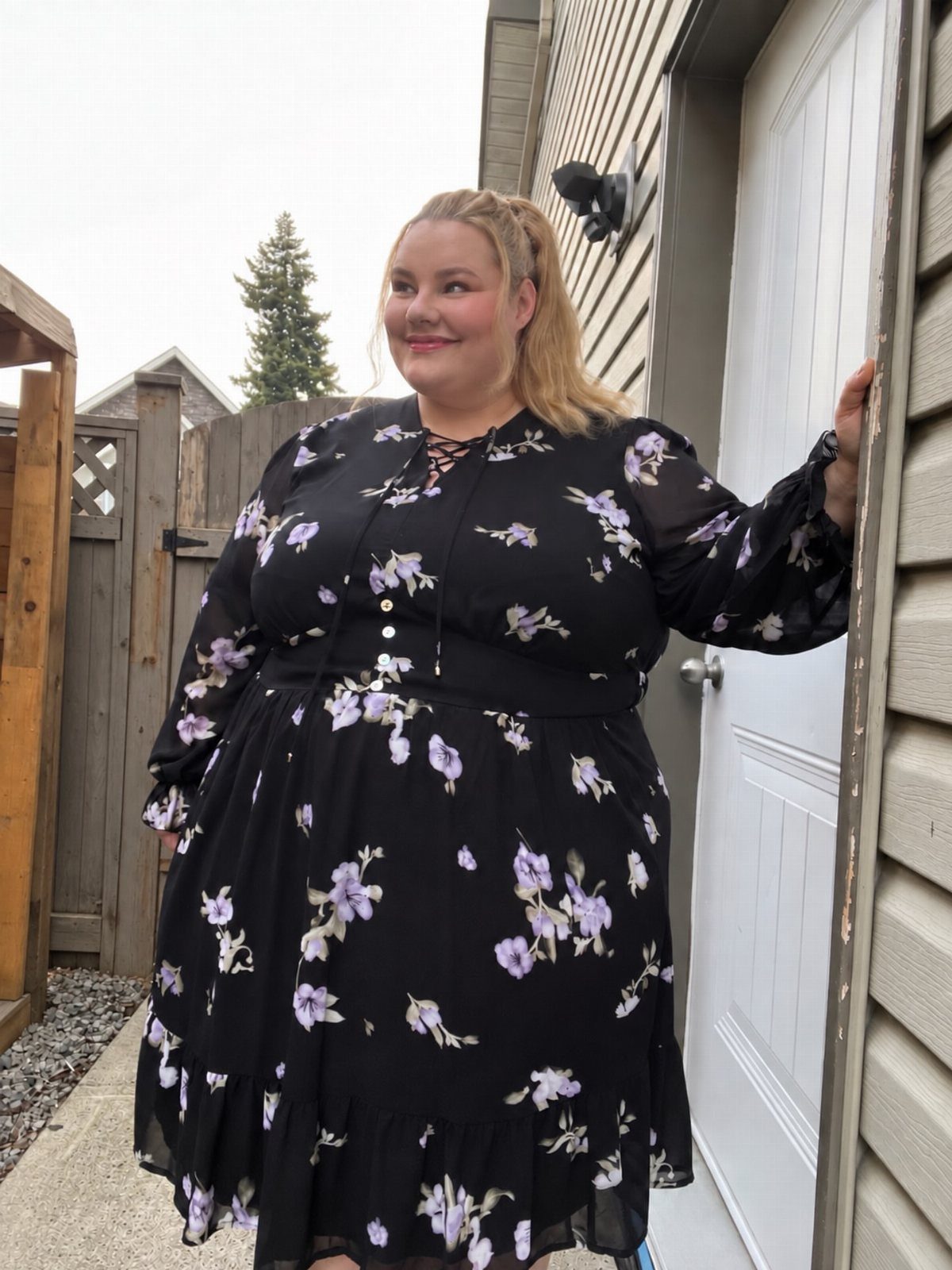 A curvy woman posing in a black floral midi dress standing by a white door.