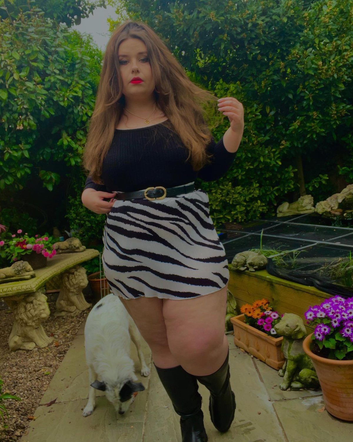 A woman in a zebra print skirt and black boots posing in a garden.