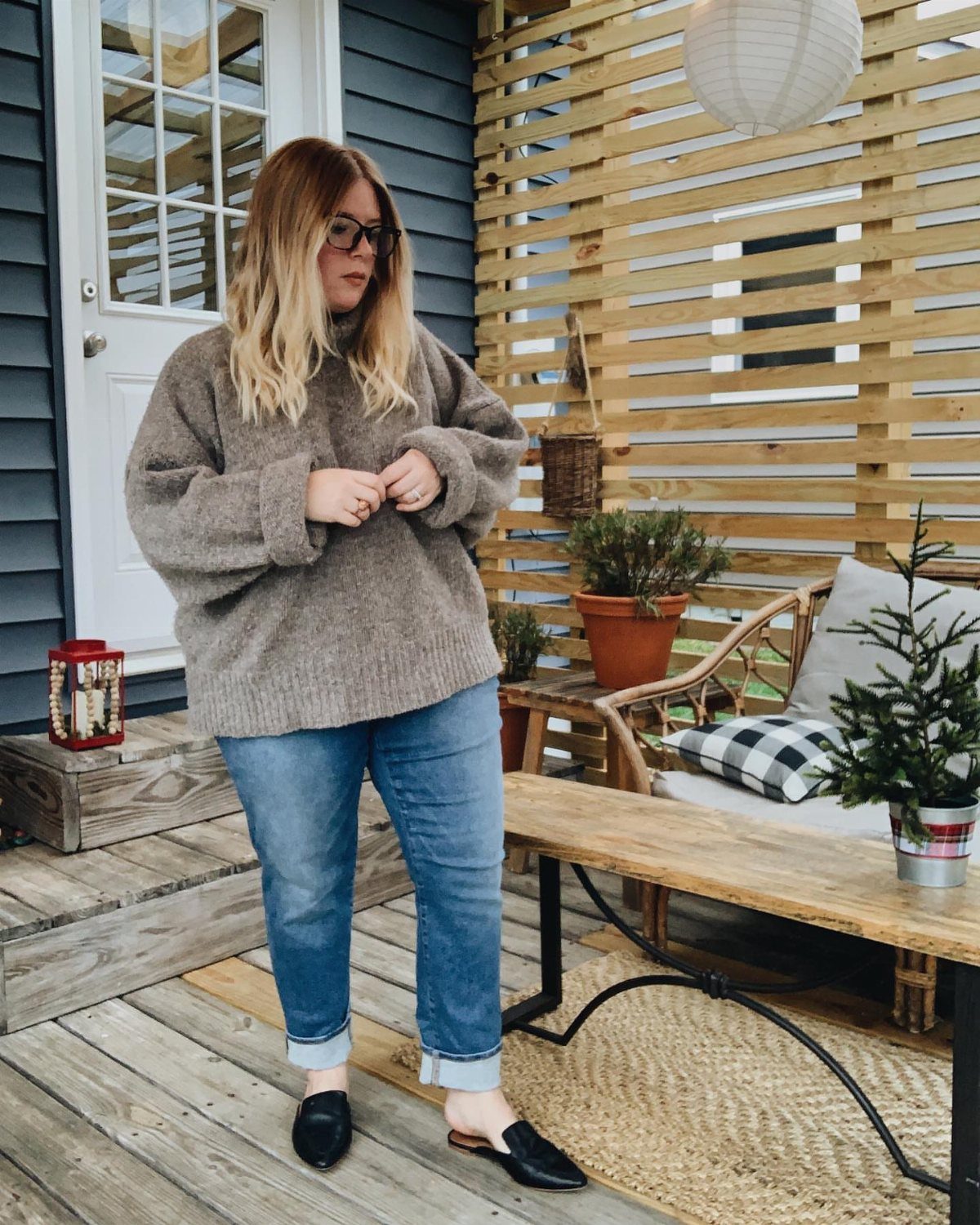 A woman wearing an oversized brown knit sweater and blue jeans standing on a porch.