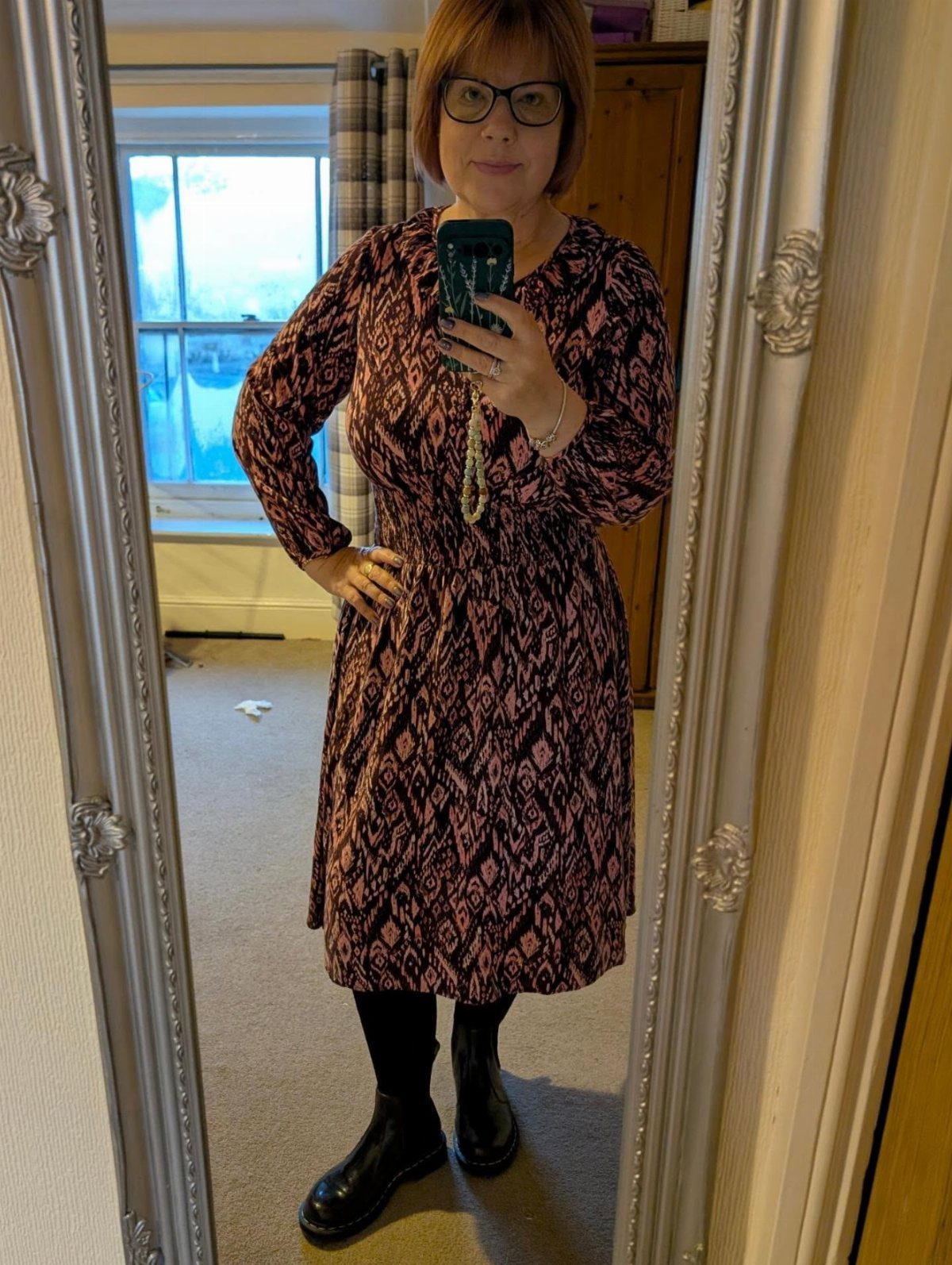 A woman posing in a patterned pink and black dress with black tights and boots.
