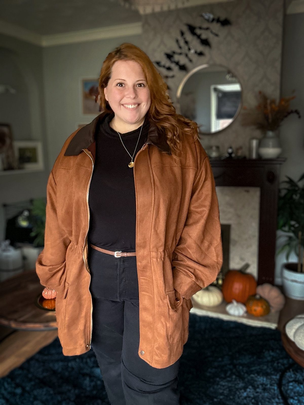 A smiling woman wearing a brown suede jacket and black turtleneck standing near pumpkins.