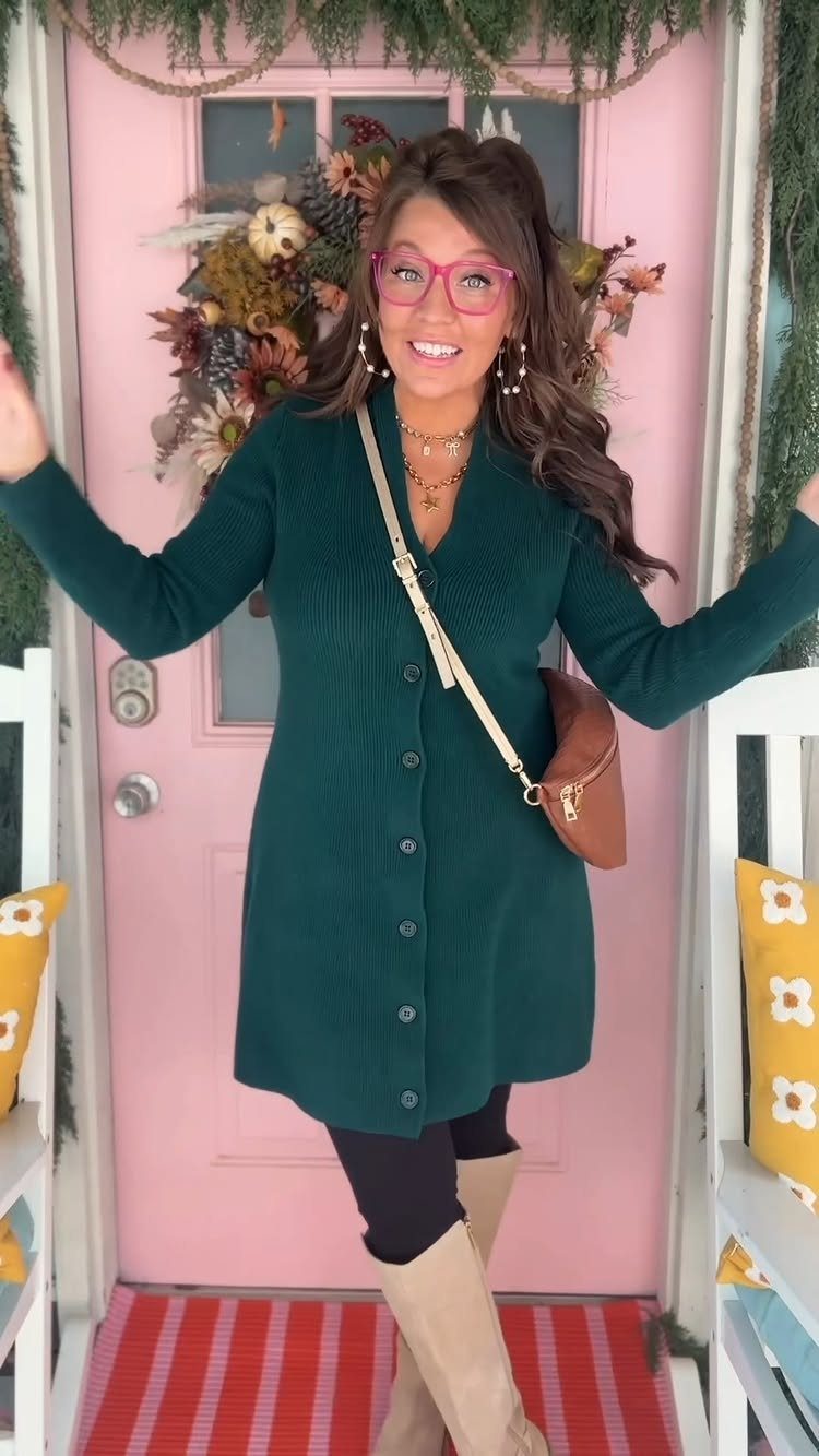 A woman wearing a green ribbed sweater dress and black leggings posing by a pink door.