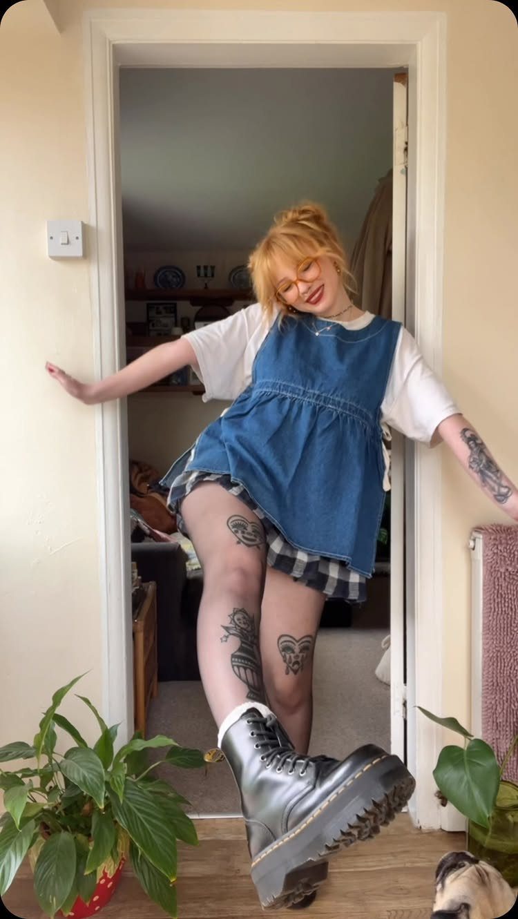 A smiling woman wearing a denim pinafore, plaid skirt, patterned tights, and black boots.