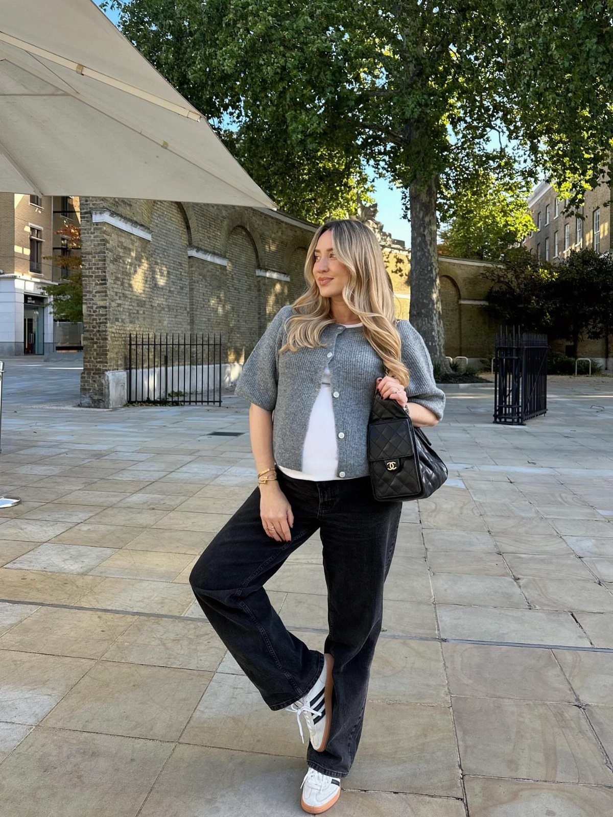 A woman wearing a grey cardigan, white top, and black jeans standing on a stone plaza.