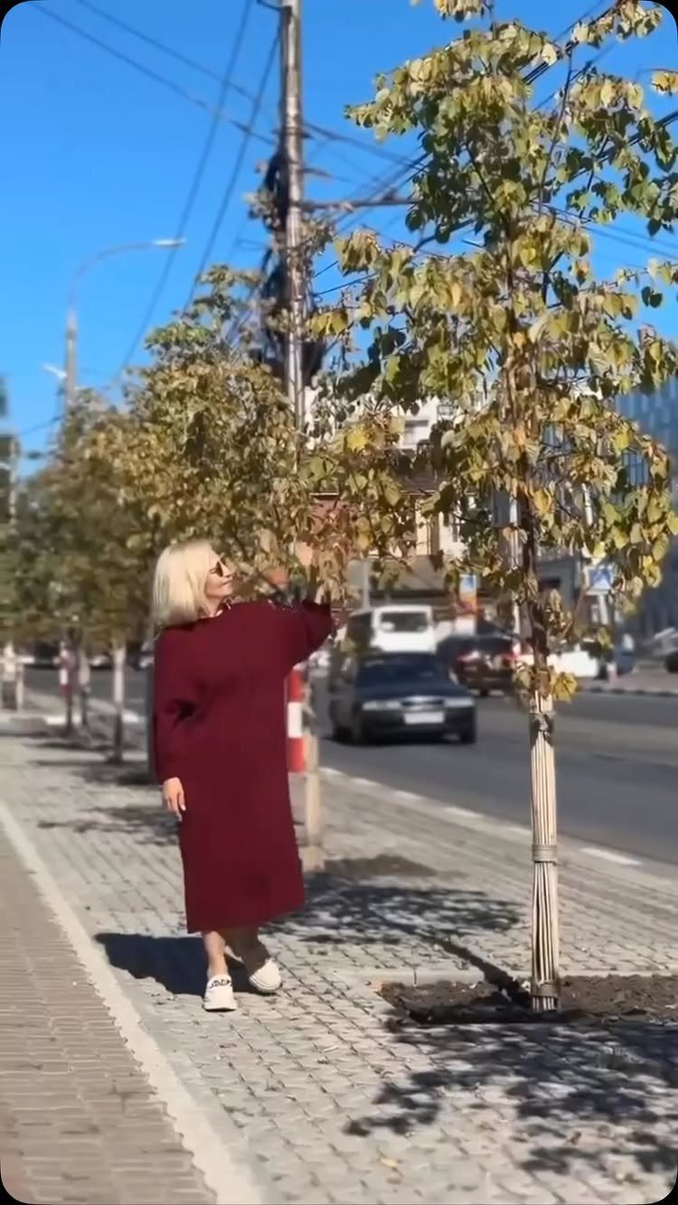 A woman in a burgundy midi dress standing next to a tree on a sunny sidewalk.