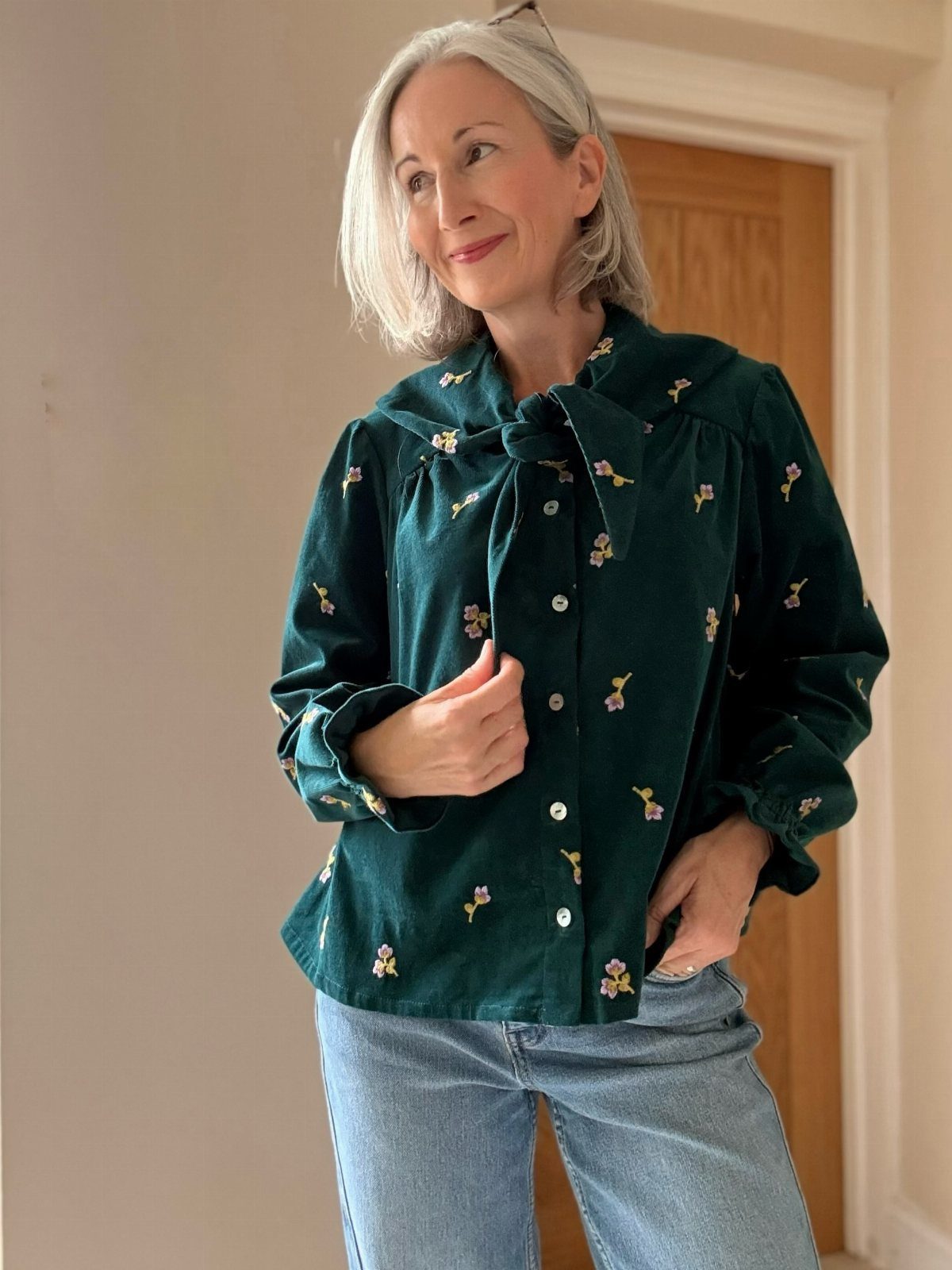 A woman smiling while wearing a dark green floral button-up shirt and blue jeans.