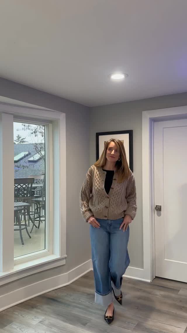 A woman posing in a tan knit cardigan, black top, and wide-leg blue jeans.