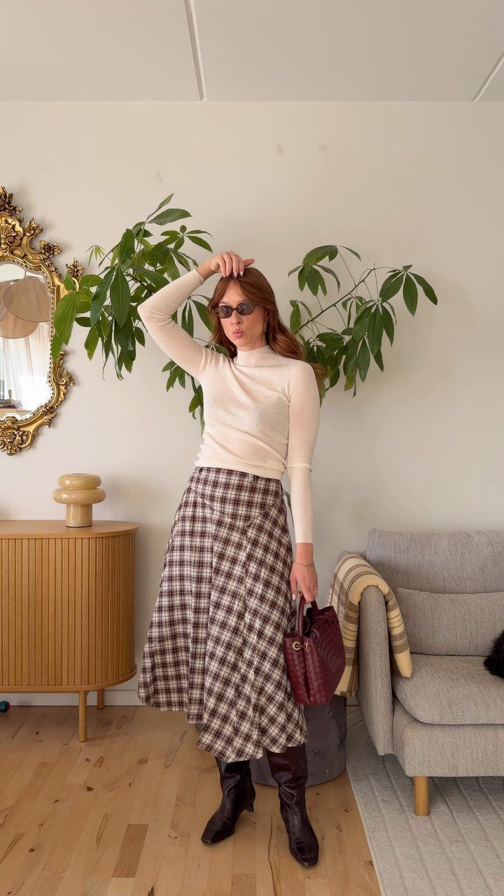 A woman posing in a cream turtleneck, plaid midi skirt, and dark brown boots.