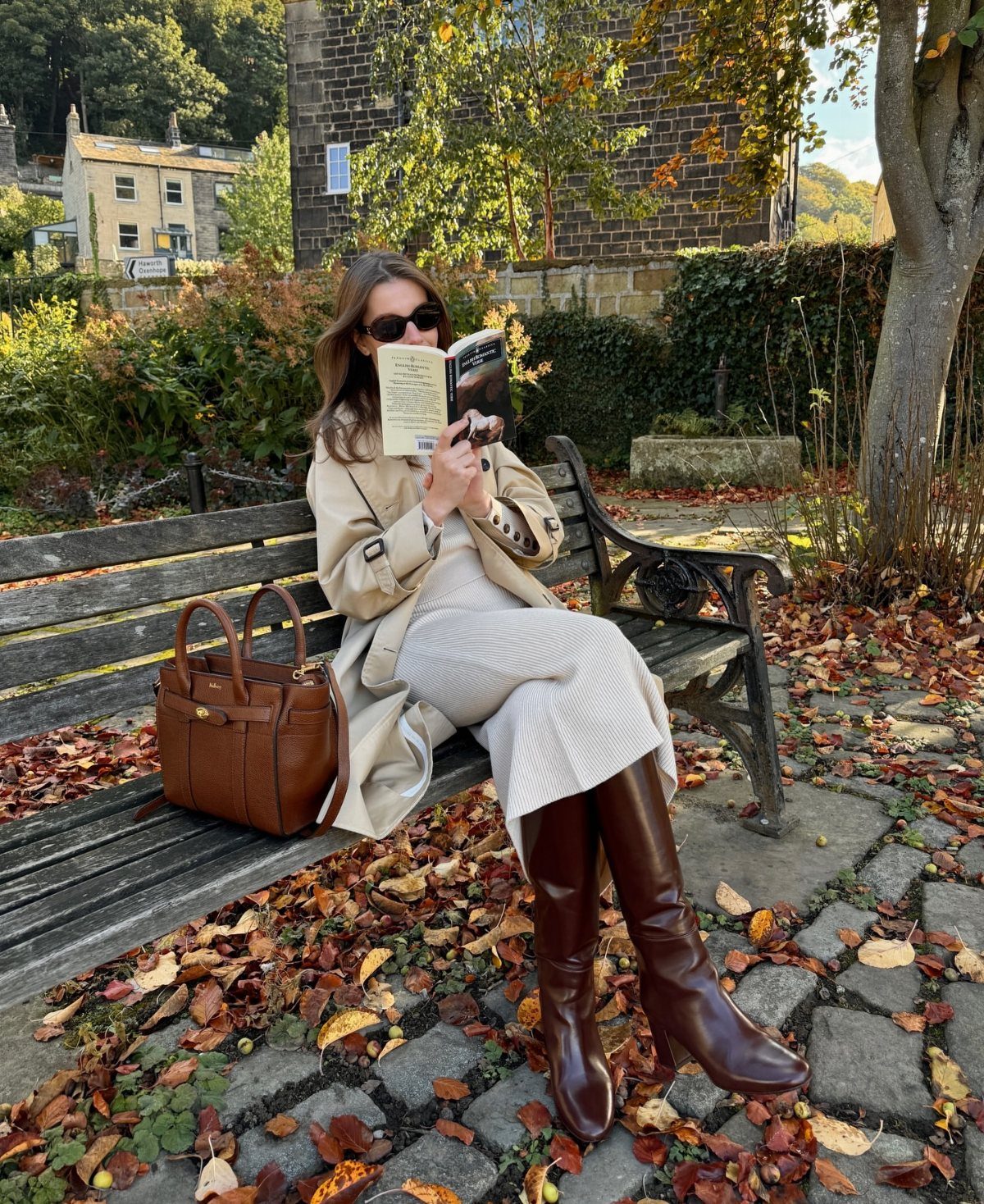 Woman in a beige trench coat and cream knit skirt sitting on a park bench reading.