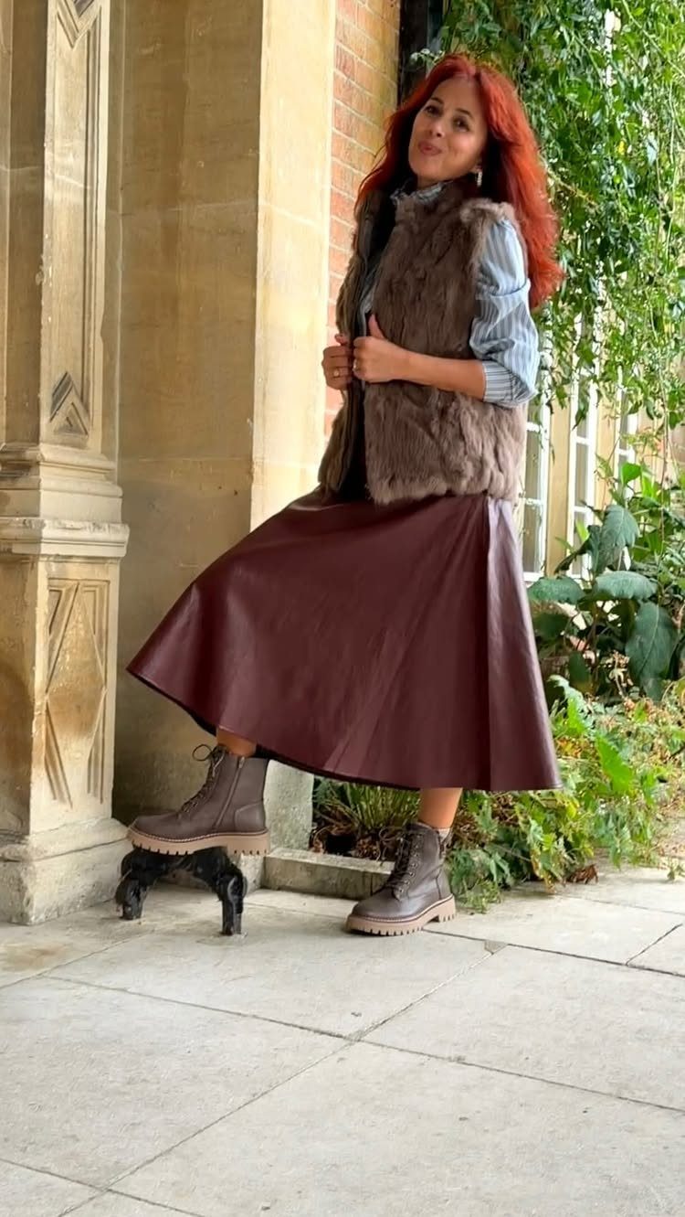 Woman with red hair wearing a fur vest, burgundy skirt, and brown ankle boots.