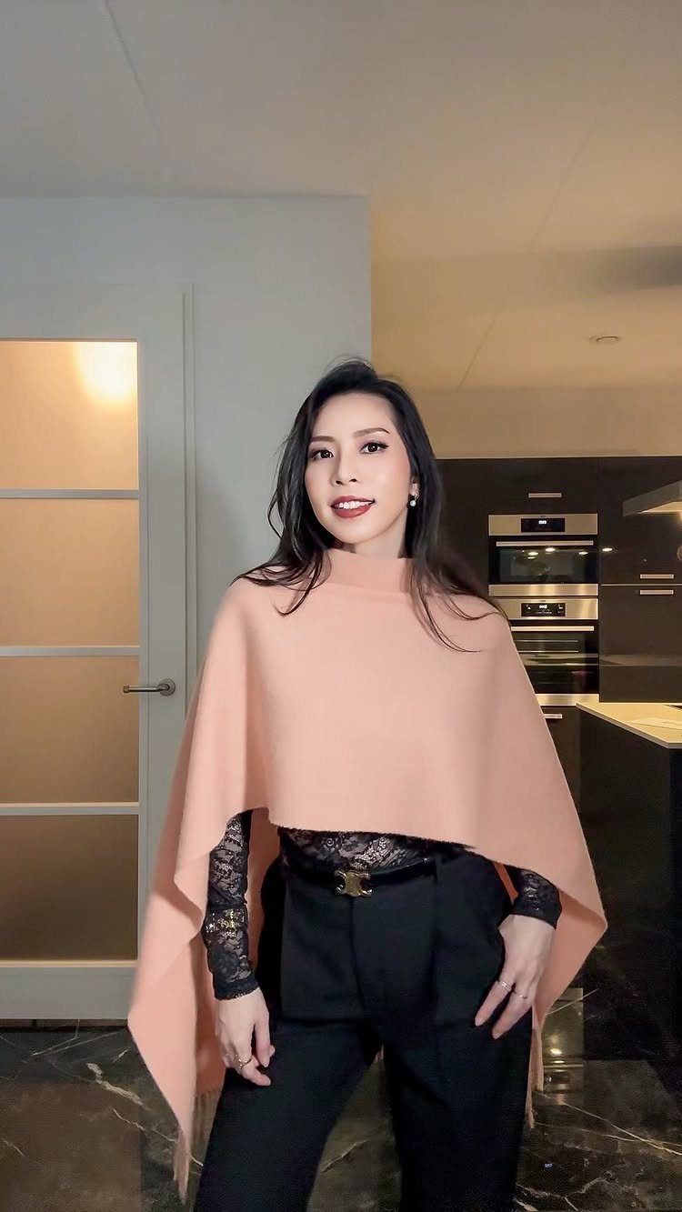 A woman posing in a peach cape, black lace top, and black trousers in a kitchen.