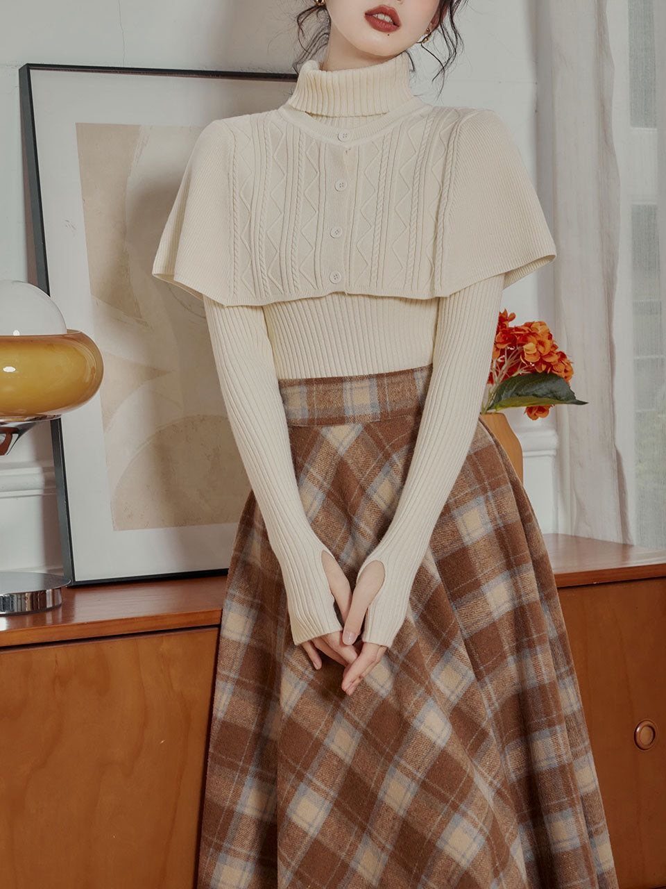 A person wearing a cream capelet turtleneck sweater and a brown plaid midi skirt.