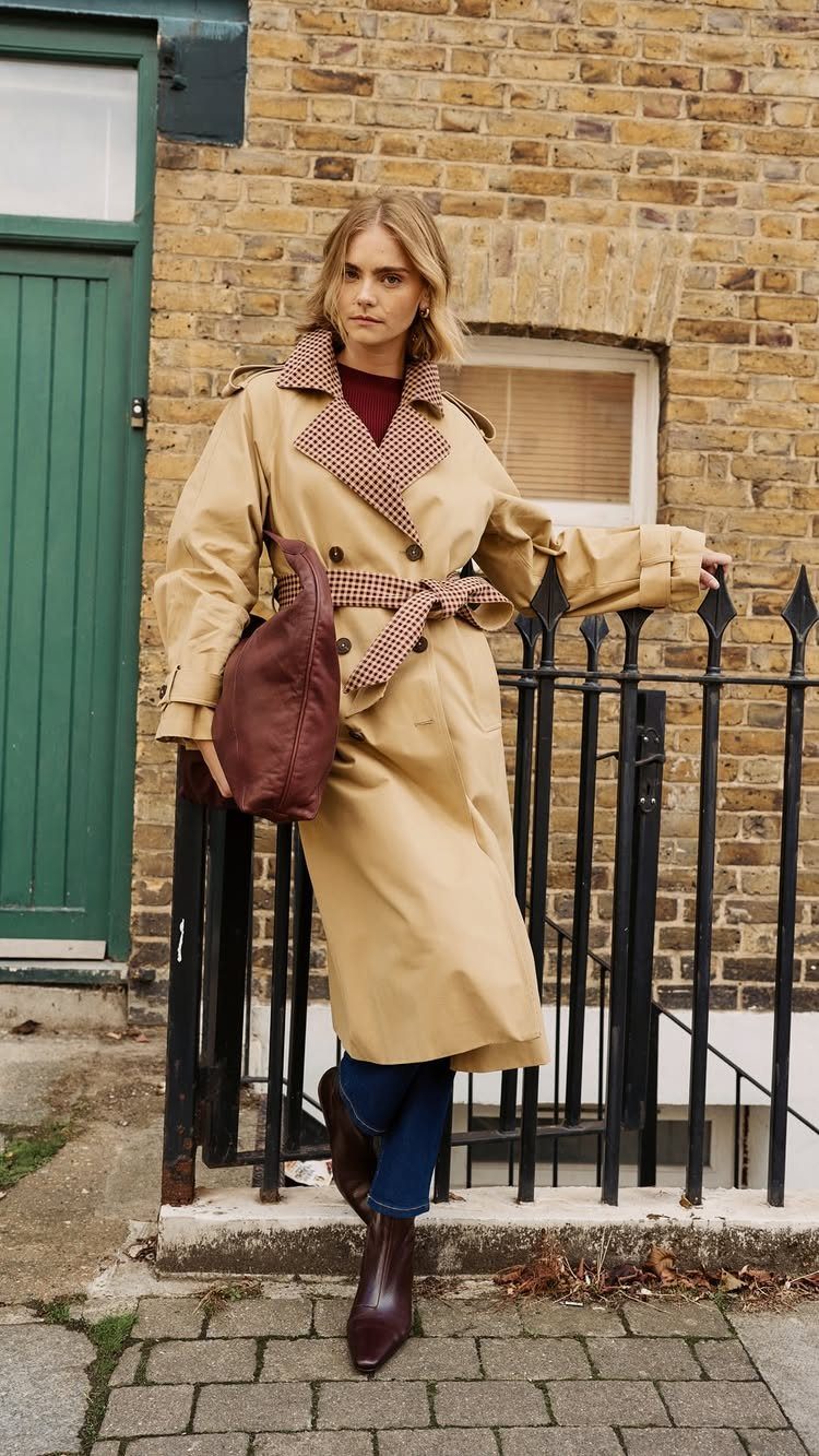 A woman wearing a long tan trench coat and dark blue jeans standing by a black fence.