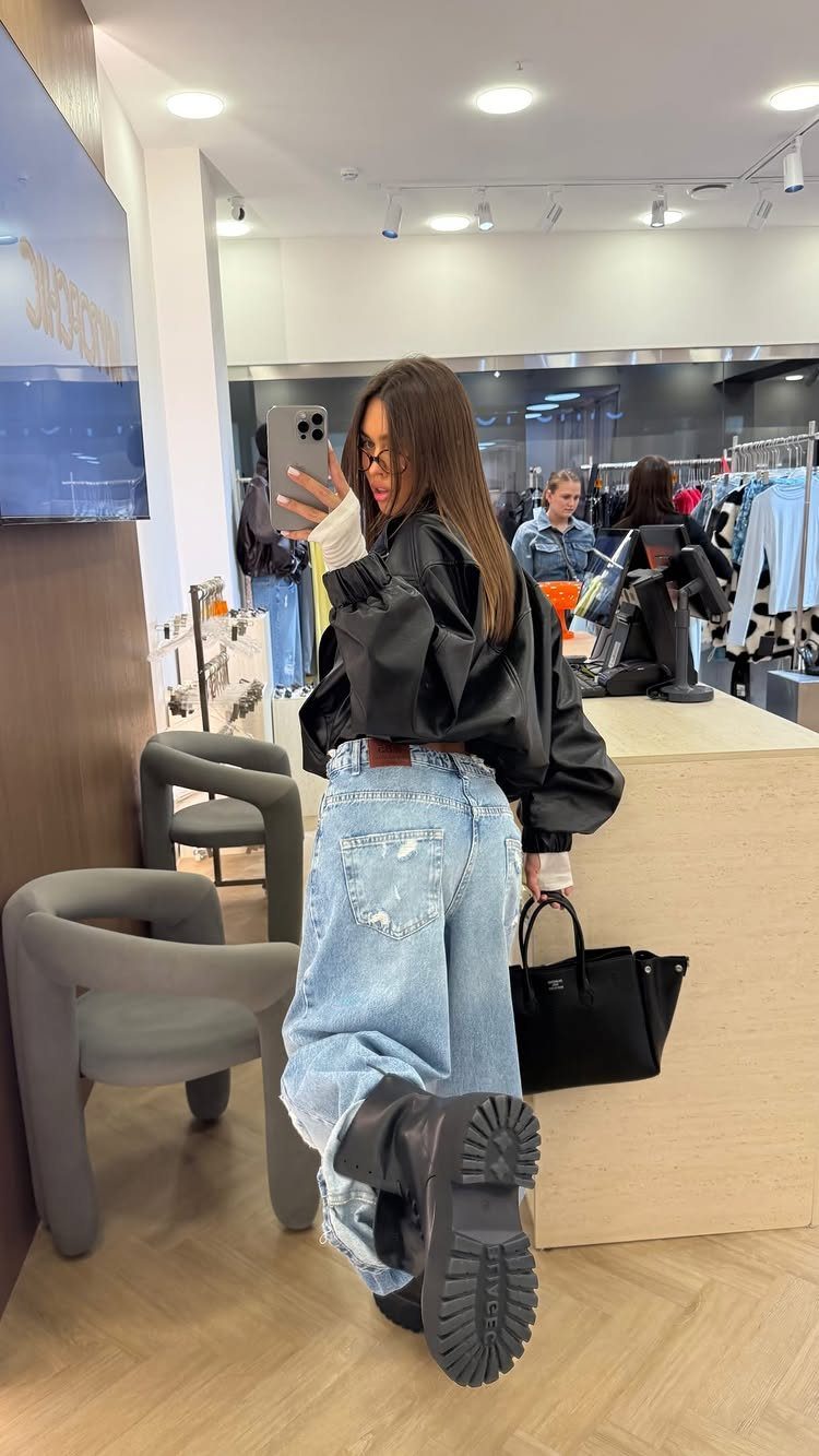 Woman in baggy denim jeans and a black leather jacket taking a mirror selfie.