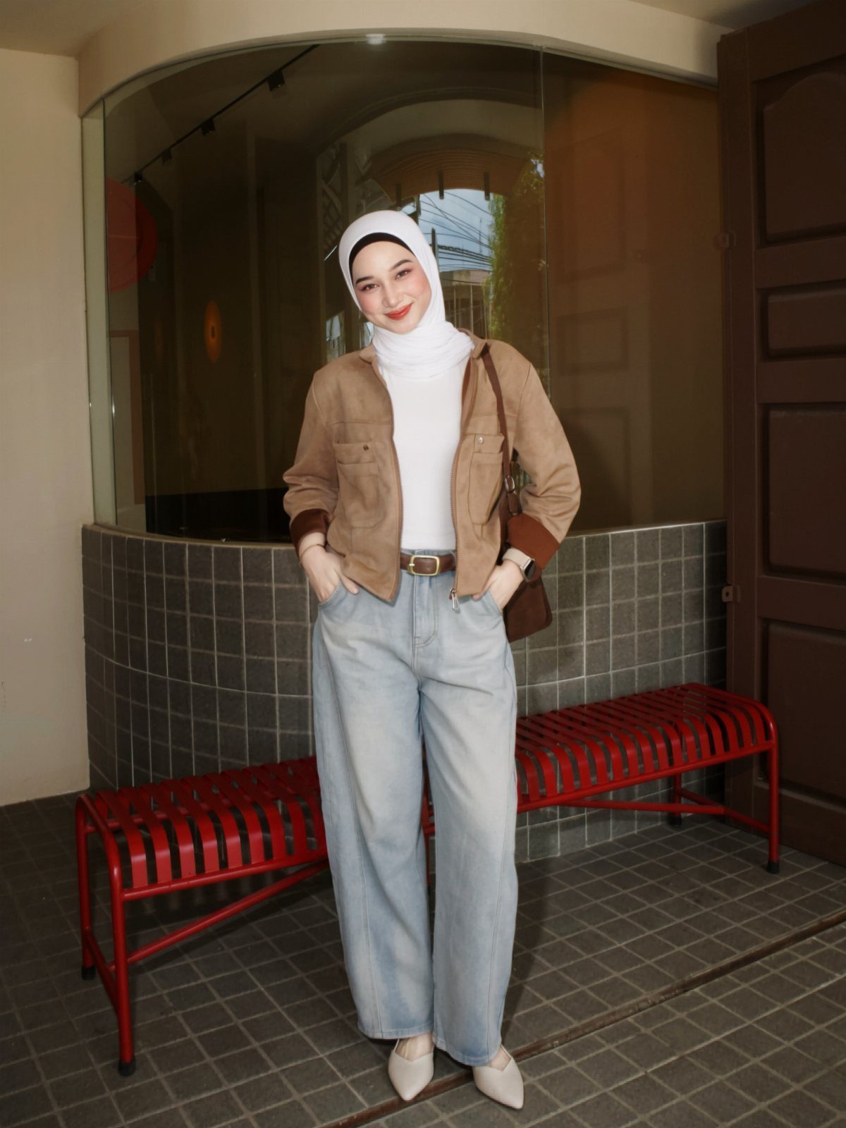 A woman in a white hijab, brown jacket, and baggy jeans standing outdoors.
