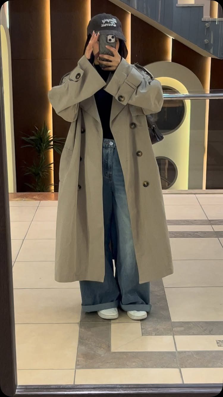 A person wearing a long beige trench coat and baggy blue jeans posing for a mirror selfie.