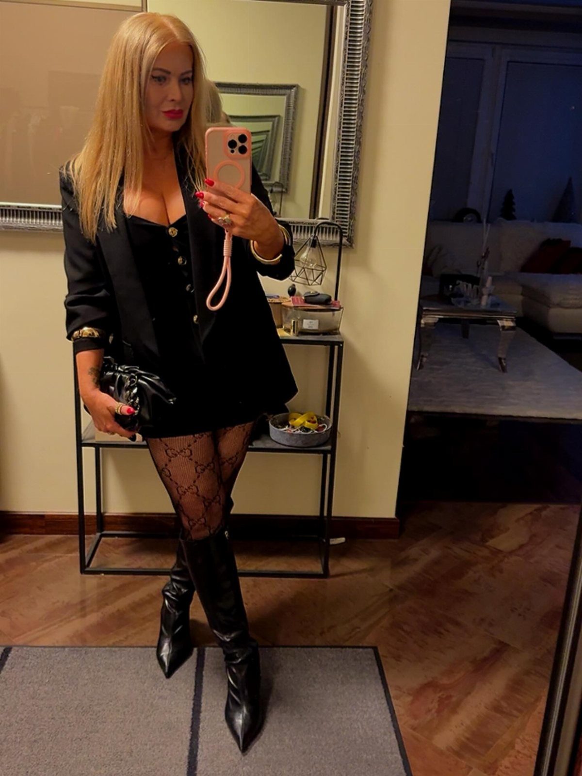 Blonde woman in a black blazer, patterned tights, and black boots posing for a mirror selfie.