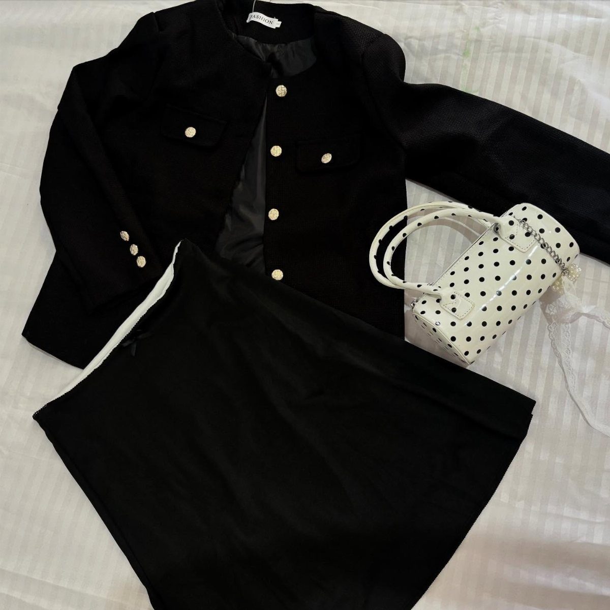 Black blazer with gold buttons, a black skirt, and a polka dot handbag on white fabric.