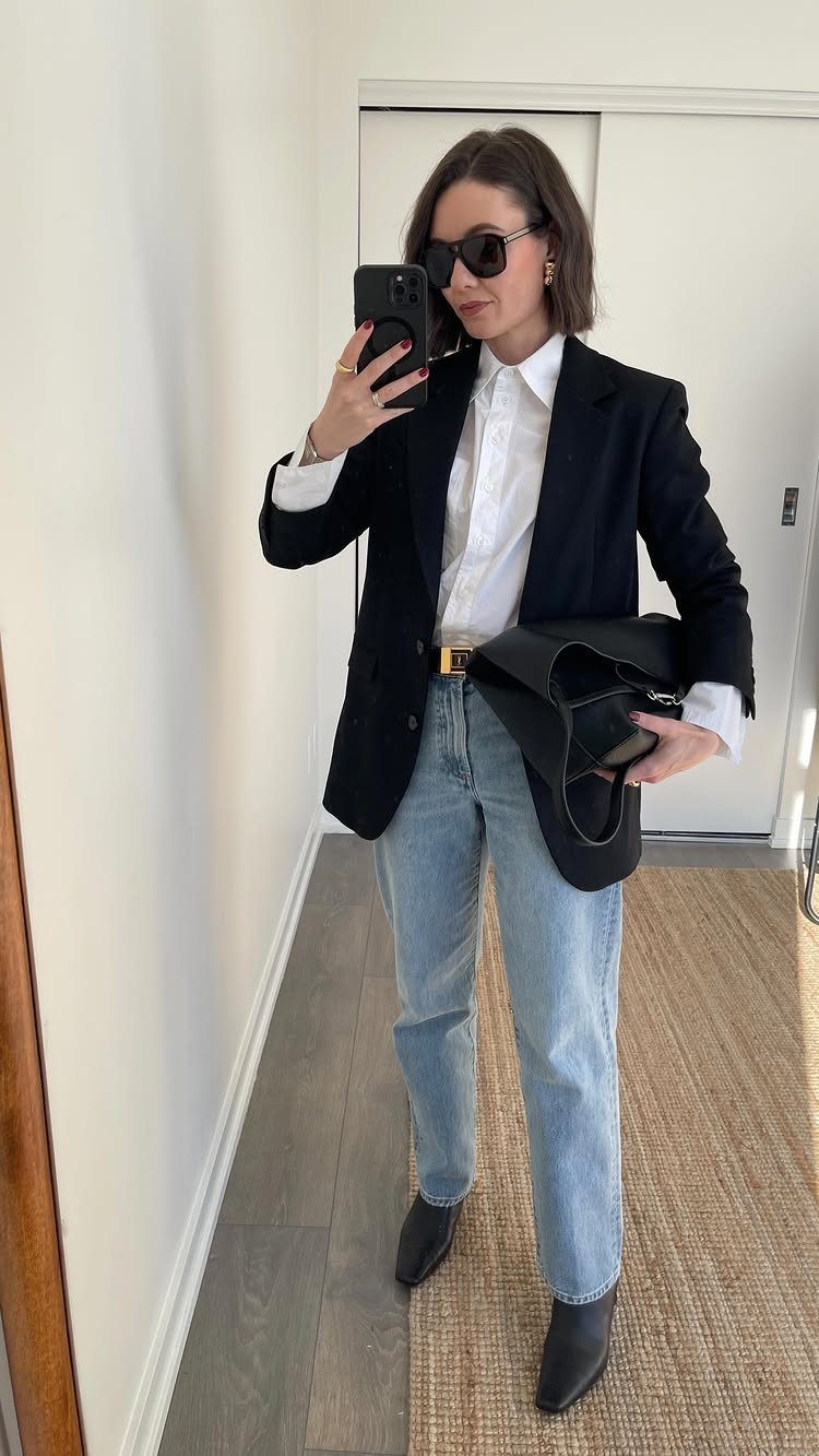 Woman in a black blazer, white shirt, and light blue jeans posing for a mirror selfie.