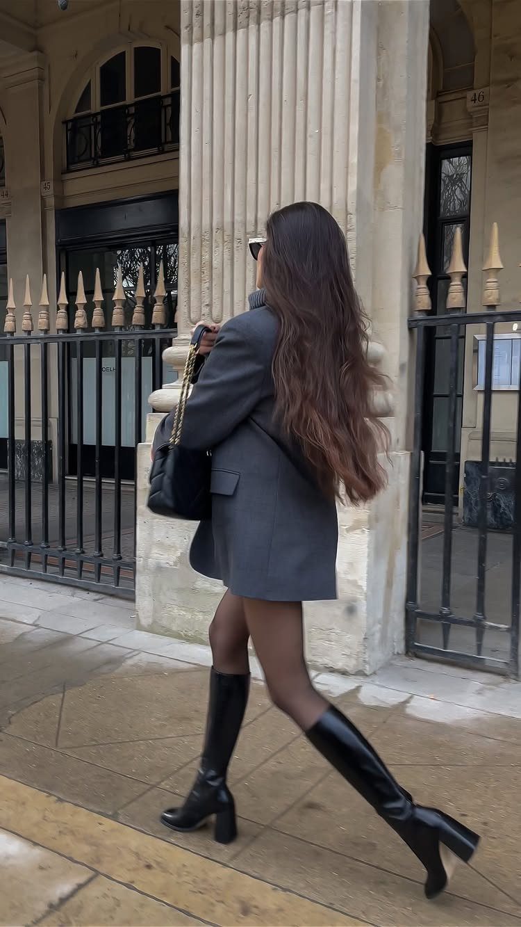 Woman walking in a grey blazer dress and tall black leather boots near a stone building.
