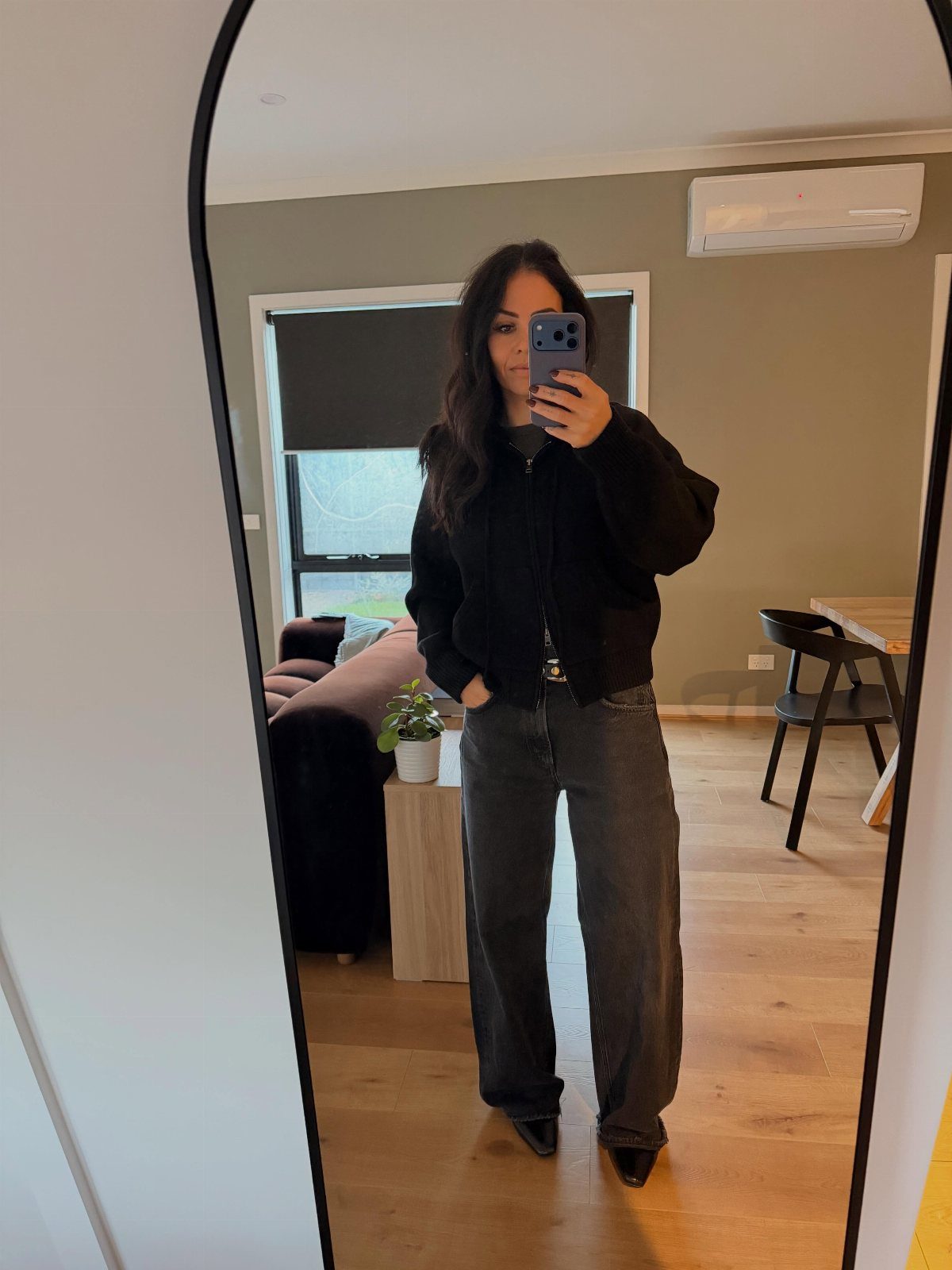 Woman in black cardigan, wide-leg jeans, and black boots posing for a mirror selfie.