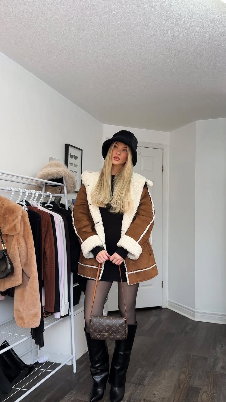 Blonde woman posing in a brown shearling jacket, black bucket hat, and black knee-high boots.
