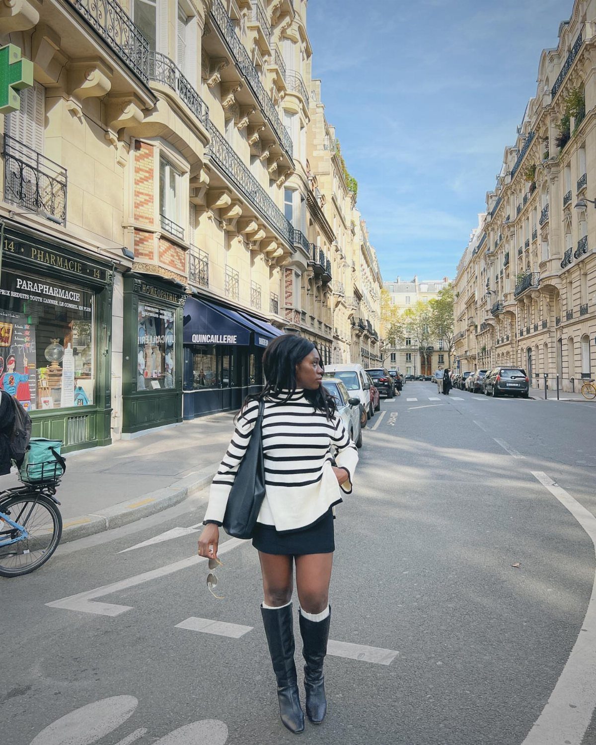 A woman in a striped top, black skirt, and tall black boots standing on a city street.