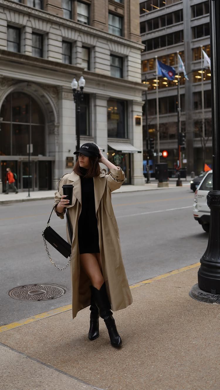 Woman in a tan trench coat, black dress, and black boots standing on a city street.