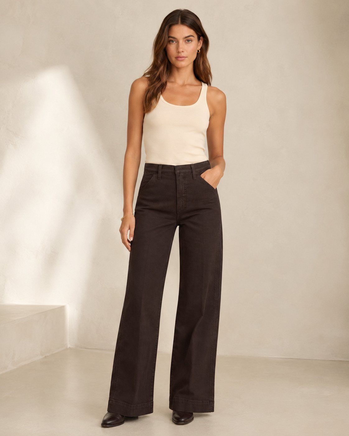 Woman wearing a beige tank top and wide-leg black jeans standing against a wall.