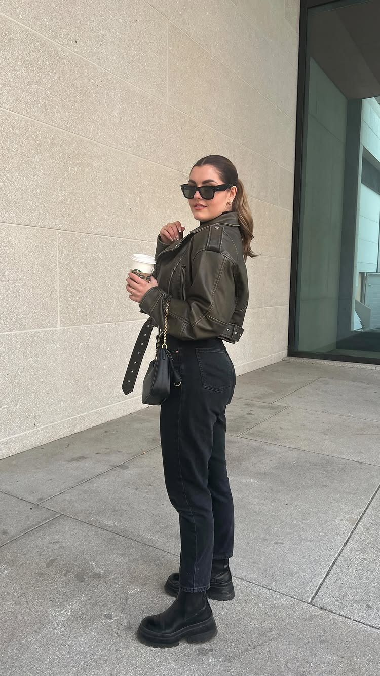 Woman in black jeans, olive leather jacket, and sunglasses holding a coffee cup outdoors.