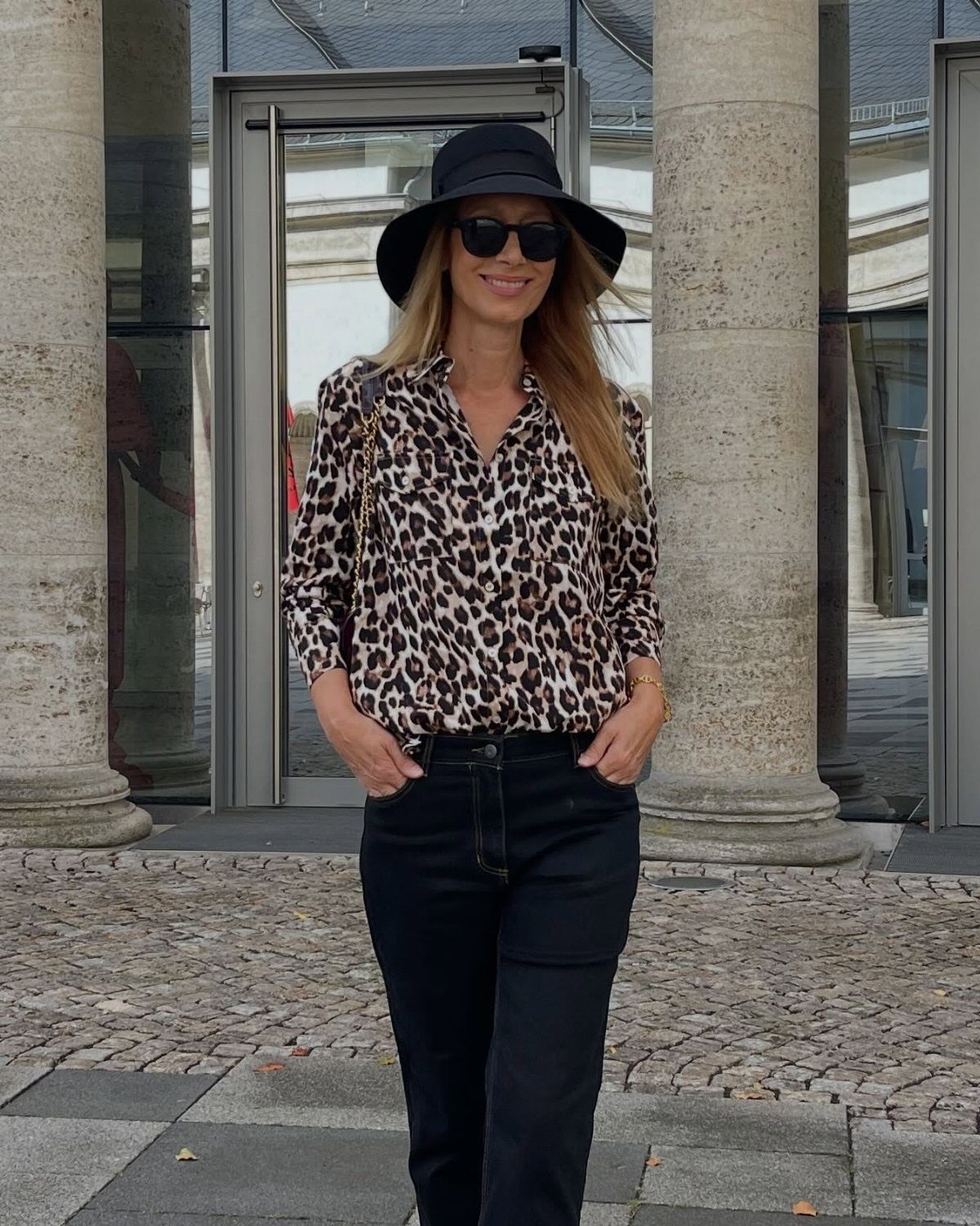 Woman wearing a leopard print blouse and black jeans with a black hat and sunglasses.