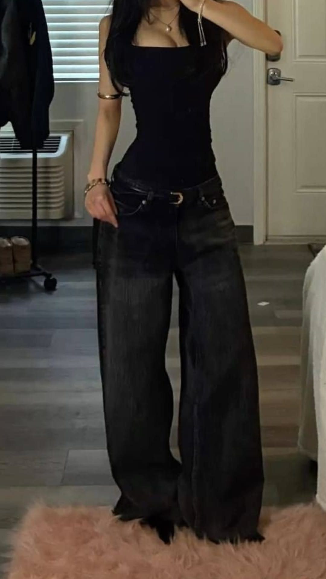 Person wearing a black tank top and wide-leg black jeans standing on a pink rug.