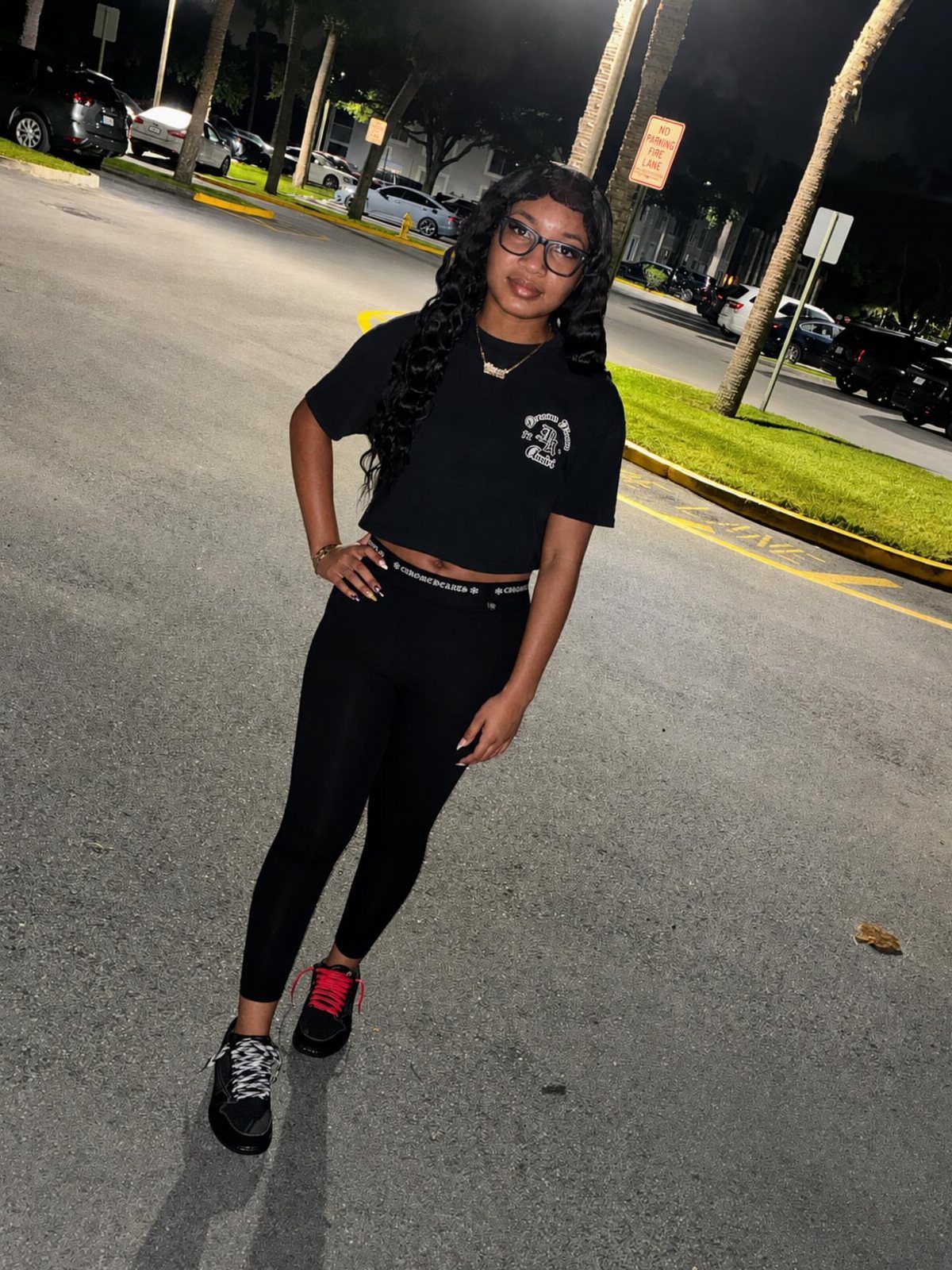 A young woman posing in a black crop top and black leggings outdoors at night.