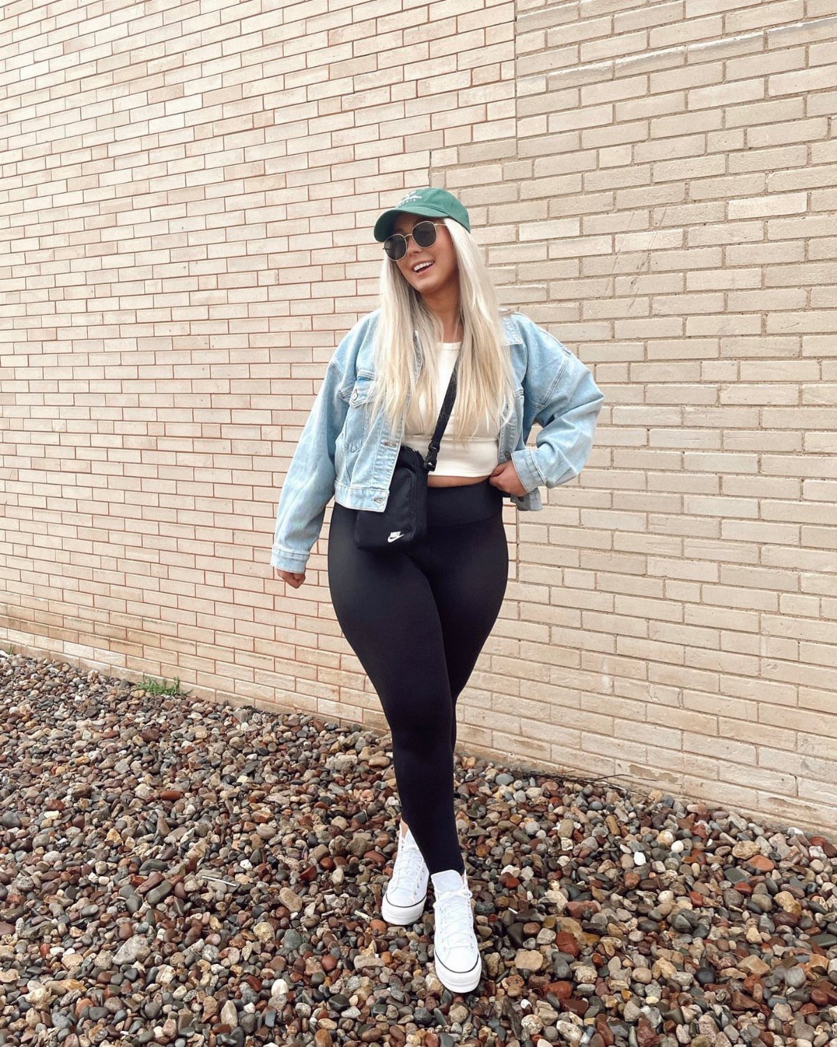 Blonde woman in a white crop top, denim jacket, and black leggings posing against a brick wall.