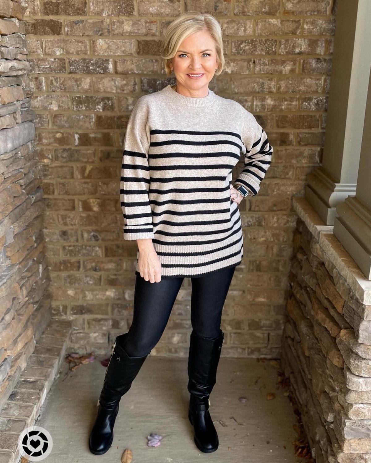Blonde woman wearing a striped long sweater, black leggings, and black boots against a brick wall.