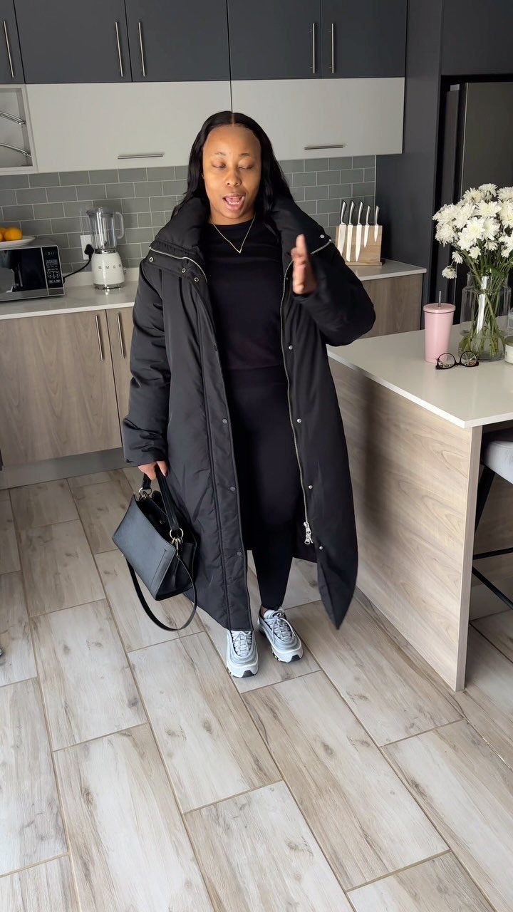Woman in black leggings, black top, and a long black hooded parka standing in a kitchen.